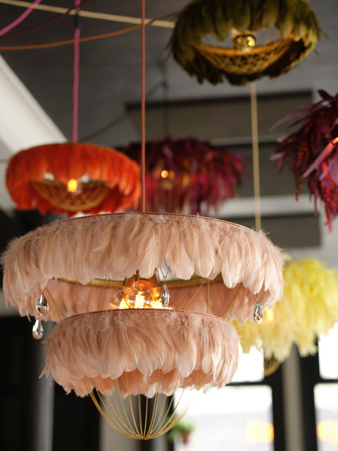 A decorative hanging chandelier with two overlapping circular tiers covered in pale blush pink feathers, with warm lights glowing from within and crystal-like hanging accents.